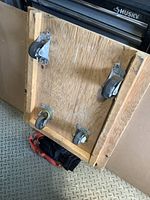 Underside of the wooden dolly showing four swivel caster wheels mounted on metal plates and red moving straps on the floor below.
