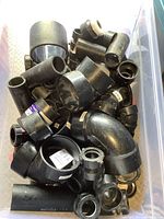 Container full of black ABS plumbing fittings including elbows, couplings, adapters, and pipe pieces piled inside a clear plastic bin.