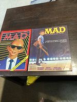 Two Mad Magazine issues from 1987 placed side by side on a dark wood surface; left one shows Alfred E. Neuman with sunglasses, right one a boy holding guitar on stage.