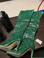 Green snakeskin knee-high boots with pointed toe and stiletto heel lying on table with other black boots nearby.