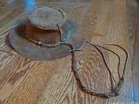Full view of the brown buckskin leather hat laid on hardwood floor showing brim, crown, and long chin strap.