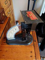 Side angle showing black metal postal scale with weighing tray and dial indicator on front marked with weight measures.