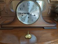 Front view of the vintage mantle clock showing the silver dial with black numbers, black clock hands, brass pendulum on wooden base.
