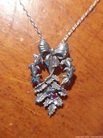 Close-up photo of the pendant showing the silver bow atop a wreath with detailed leaves and a small ruby inset.