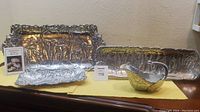Full view of the four aluminum serveware pieces displayed on a surface with a yellow cloth. Includes all trays and gravy boat with elephant motifs.