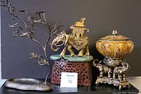 Front view of brass elephant on green stone and reddish metal base, metal jewelry display tree, painted wooden bowl with lid on elephant trio stand, ceramic plate with raised elephant head.
