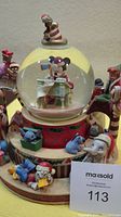 Front/side view of Disney Christmas snow globe showing Mickey Mouse inside the globe reading a newspaper, surrounded by other Disney characters like Dumbo and Stitch at the base, with Jiminy Cricket on top.