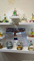 Photo showing multiple Disney mini snowglobes arranged on white shelves with certificates of authenticity behind them, showcasing various characters and themes.