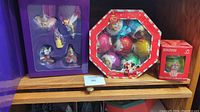 Wide view showing three unopened Disney ornament box sets on wood shelf: purple box with character figurines, octagonal box with glittery ball ornaments, and small red box with mini ornament.
