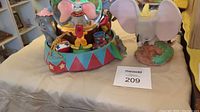 Both Dumbo circus-themed snowglobe and a separate Dumbo figure on beige cloth surface.