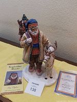 Santa figurine in traditional Laplander clothing standing next to reindeer with gift sack and decorated tree on back, along with certificate and booklet laid out on yellow cloth.