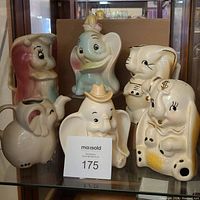 Front view of all 6 ceramic Disney Dumbo collectible items on glass shelf with brown box background.