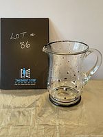 Clear glass pitcher with black and white polka dot design, shown from front angle to display shape and handle.