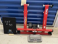 Front view of the TacX bicycle indoor trainer in red metal frame with resistance unit and gray riser block in front.