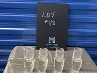 Set of four clear weighted crystal liqueur glasses arranged in a row on a grey fabric surface with black box labeled 'Lot #48' behind them.