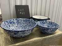 Three enamelware kitchen items displayed on a golden cloth: two blue splatter pattern bowls (large and small) and one solid blue colander with handles visible from front and side angles.