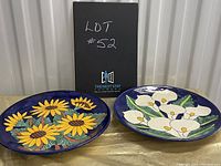 Two ceramic floral plates from Mexico - one with yellow sunflowers and one with white lilies, both on blue backgrounds.