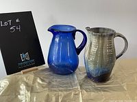 Two pitchers displayed next to each other; one blue glass and one grey/blue ceramic pottery
