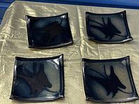 Four Japanese handmade ceramic square dishes, dark glaze with butterfly motifs, arranged on a light gold cloth.