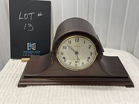Front of wooden mantle clock with curved top and round face showing Arabic numerals and clock hands.