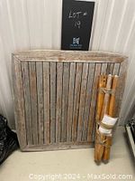 Square wooden outdoor table top with vertical slats, showing weathering and wear, leaning against wall. Four bundled wooden legs and some hardware placed adjacent to tabletop.