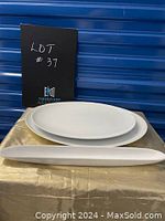 Two white serving platters displayed stacked together, showing one wider oval platter underneath and a long narrow platter in front, placed on a gold cloth with blue storage background