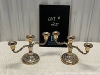 Pair of silver-plated three-arm candelabras with decorative embossed bases for taper candles, shown together with auction lot number.