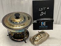 Silverplate chafing dish with glass lid and metal burner stand, alongside covered butter dish with bird handle, displayed on carpeted surface.