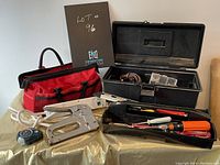 Two toolboxes side by side: a soft red fabric toolbox filled with assorted tools and a hard black plastic toolbox containing more tools including a staple gun and screwdrivers.