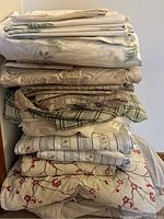 Stacked double bed linens with multiple folded sheets and pillowcases showing various floral and plaid patterns in muted colors.