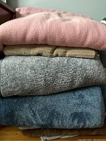 Stack of four large blankets and throws with different colors and textures including pink, beige knitted, gray fleece and dark blue plush.