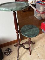 Two green marble topped wooden Bombay Company plant stands, one taller and one shorter, on tile floor near wall