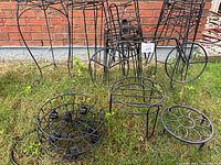 Overview photo showing entire set of black metal outdoor plant stands including bicycle-shaped stand, circular rotating holder, and hooks on grass in front of brick wall.
