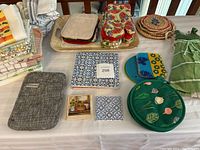 Full view of the kitchen items lot showing stacked tea towels, oven mitts, hot pads, KitchenAid pocket, and decorative ceramic tiles on a table.