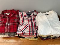 Folded stack of assorted ladies button-up shirts in various patterns and solid colors, laid on a wooden surface.