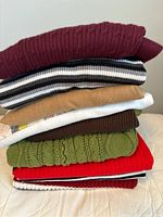 Stack of 8 folded sweaters showing variety of colors and knit patterns including solid burgundy, striped, camel, brown, green cable knit, red, and more.