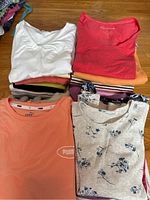 Multiple stacks of folded ladies short sleeve tops of various colors and prints on a wooden floor including solid, floral, striped, and branded pieces.