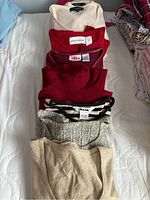 Six folded ladies short sleeve sweaters in assorted colors and patterns on a white bedspread.