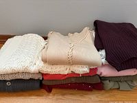 Overview of folded sweaters showing assorted colors and knit styles including beige ribbed knit with lace trim, cream knit, maroon ribbed, grey, red, pink, and olive green sweaters.