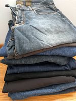 Stack of seven men's jeans folded on a wooden floor, showing different colors and textures including light faded blue, classic blue denim, dark blue, and black.