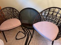 Two wicker chairs with pinkish beige cushions, disconnected table parts and round glass tabletop leaning against wall.
