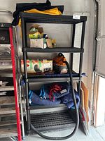 Photo showing black resin 5-shelf storage unit with various garage items stored on shelves and floor space in front.