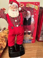 Photo of the red suspenders, plaid shirt Santa figurine standing on a black base, with packaging in background showing another Santa.