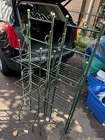 Photo showing two free standing folding metal shelves with decorative scroll tops and spherical finials, placed outdoors near a vehicle and other items.