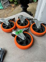 Four heavy duty locking caster wheels with orange rubber tires and metal brackets arranged on a surface.