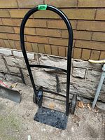 Photo of a black metal moving dolly with two wheels and an arched handle, resting outdoors against a stone wall.