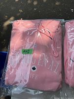 Front view of pink hoodie sweatshirt in original plastic packaging showing partial white printed text and maple leaf graphic.