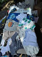 Overview photo showing variety of new with tags baby and kids clothes spread on a table in outdoor setting.
