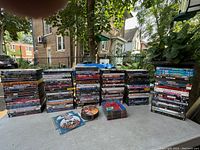Photo of multiple stacks of original DVD movie cases outdoors, showing a large quantity of varied movie titles.