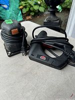 Photo shows a black palm sander with an orange switch and the power cord coiled next to a black and red Lonsdale electric stapler with its power cord wrapped around it, placed on a light-colored surface outdoors.